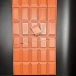 May include: A large, rectangular chocolate bar with a broken piece missing. The bar is made of individual squares and is a deep orange colour.