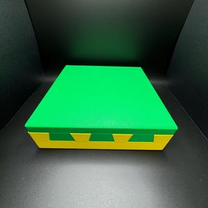 May include: A green and yellow box with a lid. The box is made of a smooth, hard material. The lid is slightly raised above the box.