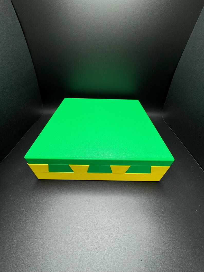 May include: A green and yellow box with a lid. The box is made of a smooth, hard material. The lid is slightly raised above the box.