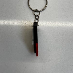 May include: A keychain with a silver ring and a rectangular charm. The charm is black, white, and red. The keychain is likely made of metal and plastic. The design is simple and modern, suitable for keys or as a decorative accessory.
