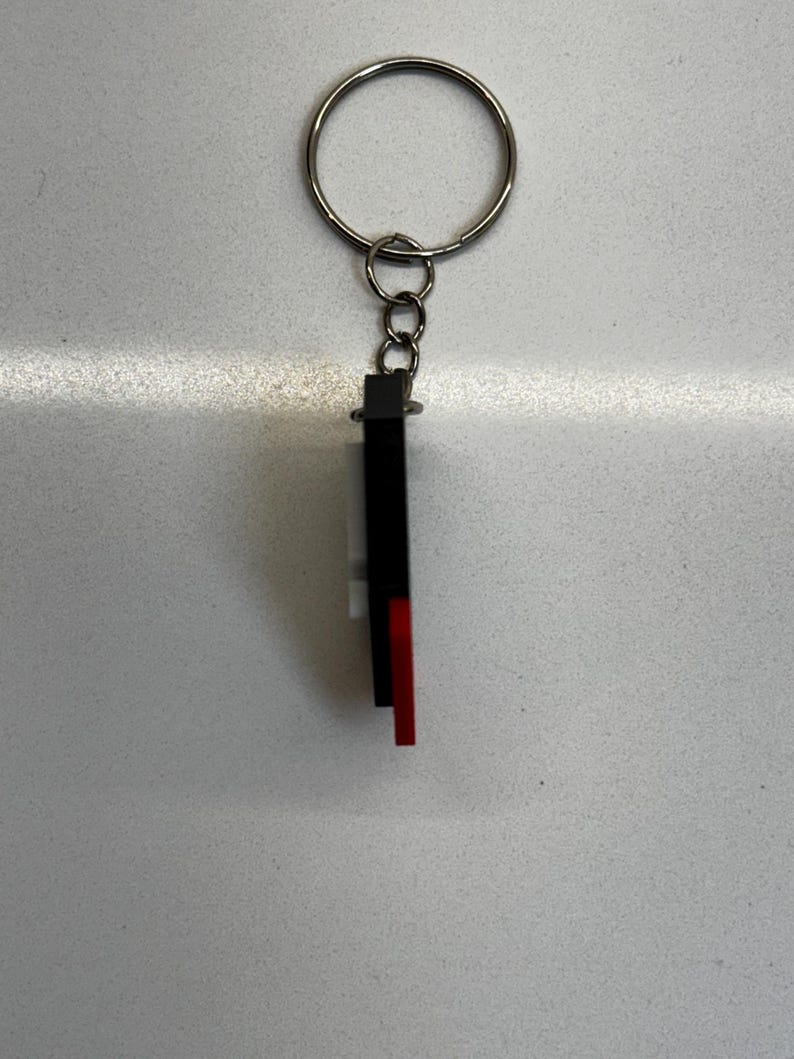 May include: A keychain with a silver ring and a rectangular charm. The charm is black, white, and red. The keychain is likely made of metal and plastic. The design is simple and modern, suitable for keys or as a decorative accessory.