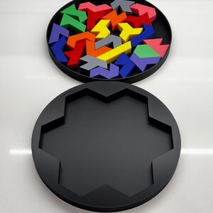 May include: A black circular puzzle box with a lid. The lid is open and contains a variety of colorful arrow-shaped puzzle pieces. The pieces are arranged in a circular pattern.