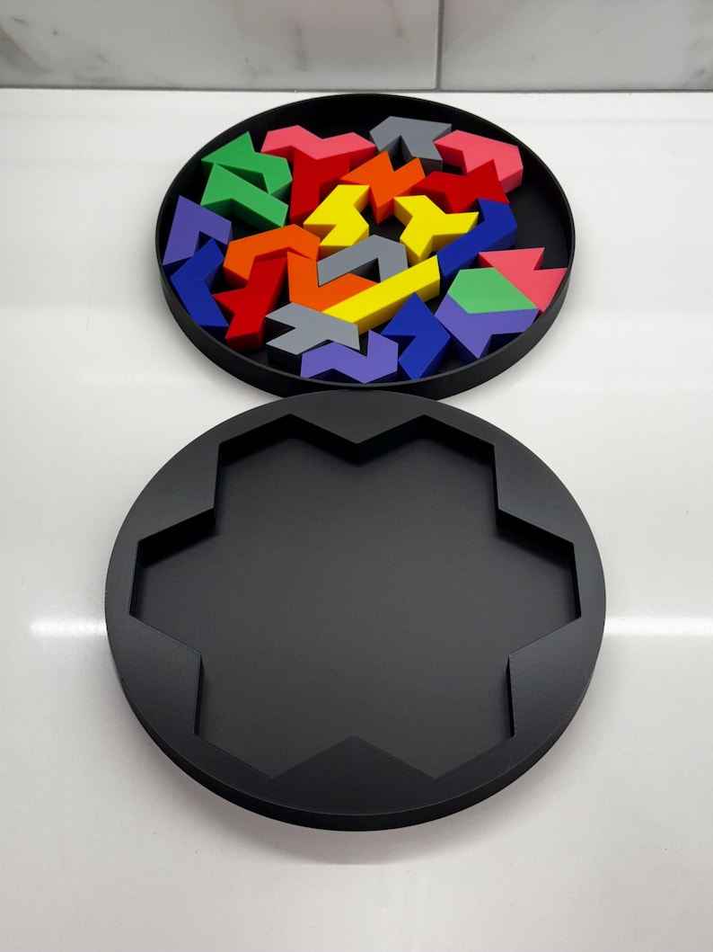 May include: A black circular puzzle box with a lid. The lid is open and contains a variety of colorful arrow-shaped puzzle pieces. The pieces are arranged in a circular pattern.