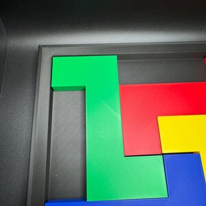 Op de afbeelding: Een kleurrijk puzzelspel met geometrische vormen in groen, rood, geel en blauw. De stukken zijn gerangschikt in een zwarte bak, met een groen L-vormig stuk prominent aanwezig. De puzzel is ontworpen voor ruimtelijk redeneren en probleemoplossing.