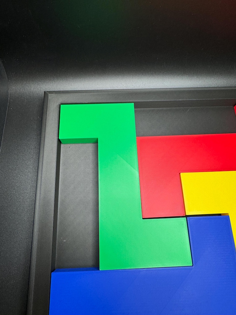 Op de afbeelding: Een kleurrijk puzzelspel met geometrische vormen in groen, rood, geel en blauw. De stukken zijn gerangschikt in een zwarte bak, met een groen L-vormig stuk prominent aanwezig. De puzzel is ontworpen voor ruimtelijk redeneren en probleemoplossing.