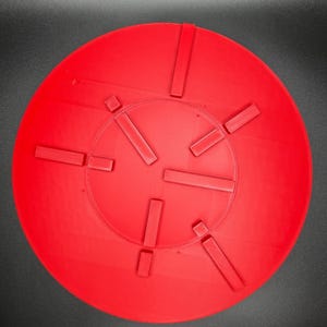 May include: A bright red, circular object with a series of rectangular protrusions evenly spaced around its perimeter. The object has a slightly textured surface and appears to be made of a solid material. The design suggests a functional or decorative item.