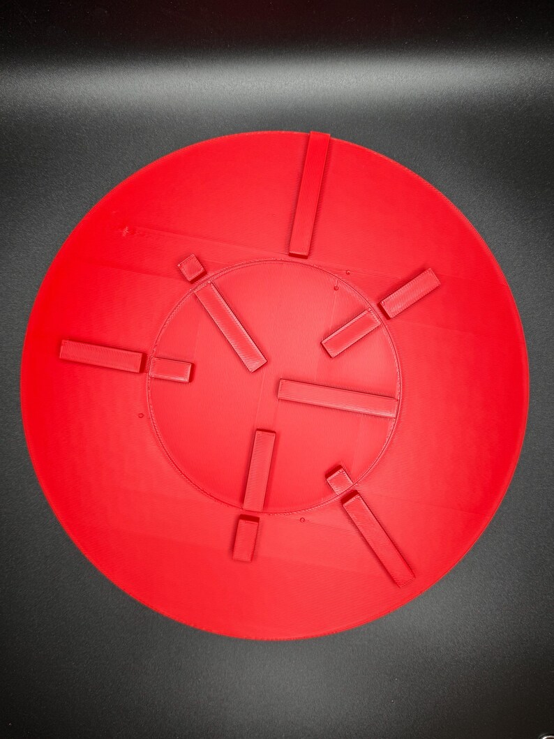 May include: A bright red, circular object with a series of rectangular protrusions evenly spaced around its perimeter. The object has a slightly textured surface and appears to be made of a solid material. The design suggests a functional or decorative item.