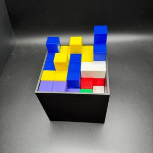 May include: A black square container filled with colorful, stacked blocks. The blocks are various colors, including blue, yellow, red, white, green, and purple. The blocks are arranged in a puzzle-like configuration.