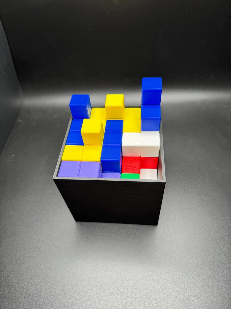 May include: A black square container filled with colorful, stacked blocks. The blocks are various colors, including blue, yellow, red, white, green, and purple. The blocks are arranged in a puzzle-like configuration.
