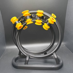 May include: A black, 3D-printed sculpture with concentric rings and a base. Yellow and red cylindrical elements are spaced around the rings. The sculpture has a modern, geometric design. The measurements are in centimetres.