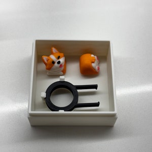 May include: A white box containing a black plastic keycap, a white and orange corgi head keycap, and a white and orange corgi tail keycap.