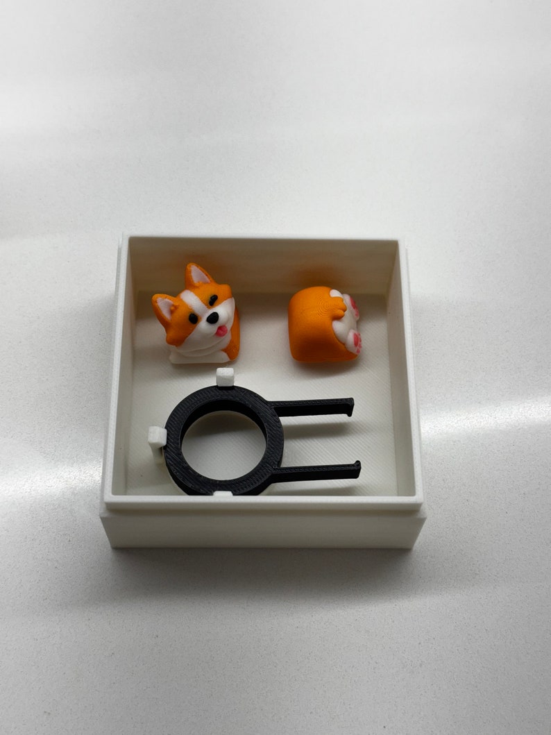 May include: A white box containing a black plastic keycap, a white and orange corgi head keycap, and a white and orange corgi tail keycap.