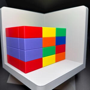 May include: A rectangular block structure composed of multiple smaller, solid-colored bricks in red, purple, yellow, green, blue, and orange. The bricks are arranged in a grid pattern and are positioned inside a white, L-shaped corner display stand.