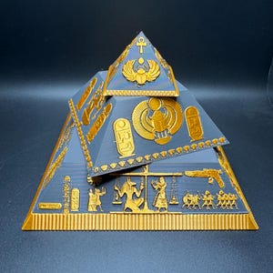 Puede incluir: Un juego de dos pir&aacute;mides decorativas con detalles dorados y grises. Las pir&aacute;mides presentan tallas jerogl&iacute;ficas, escarabajos y otros dise&ntilde;os de tem&aacute;tica egipcia. El conjunto es probablemente un art&iacute;culo decorativo o de colecci&oacute;n.