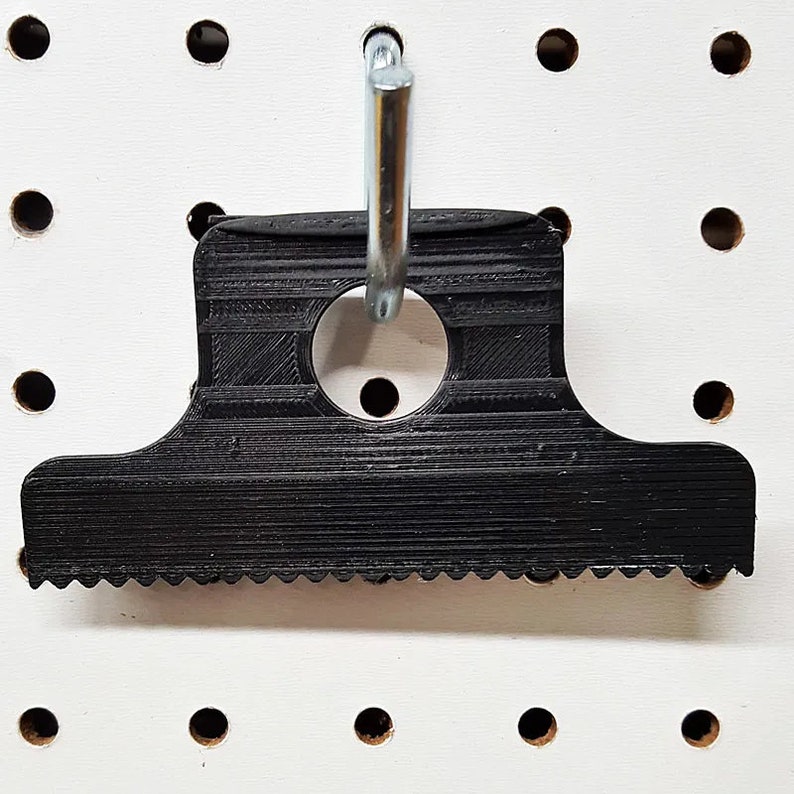 May include: Black plastic scraper tool with a serrated edge and a hole for hanging. The tool is hanging on a pegboard.