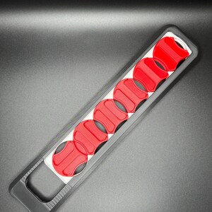 May include: A black plastic tool with a handle and eight red circular slots. The tool is designed to hold and organize small round objects.