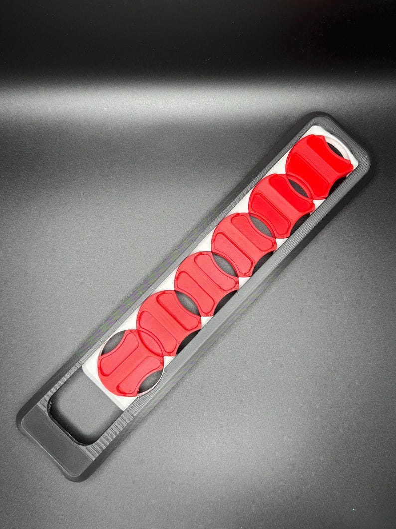 May include: A black plastic tool with a handle and eight red circular slots. The tool is designed to hold and organize small round objects.