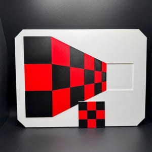 May include: A white rectangular art piece with a geometric design in red and black checkerboard pattern. A smaller, matching checkerboard square is in front of the larger piece. The artwork has a modern, abstract aesthetic.