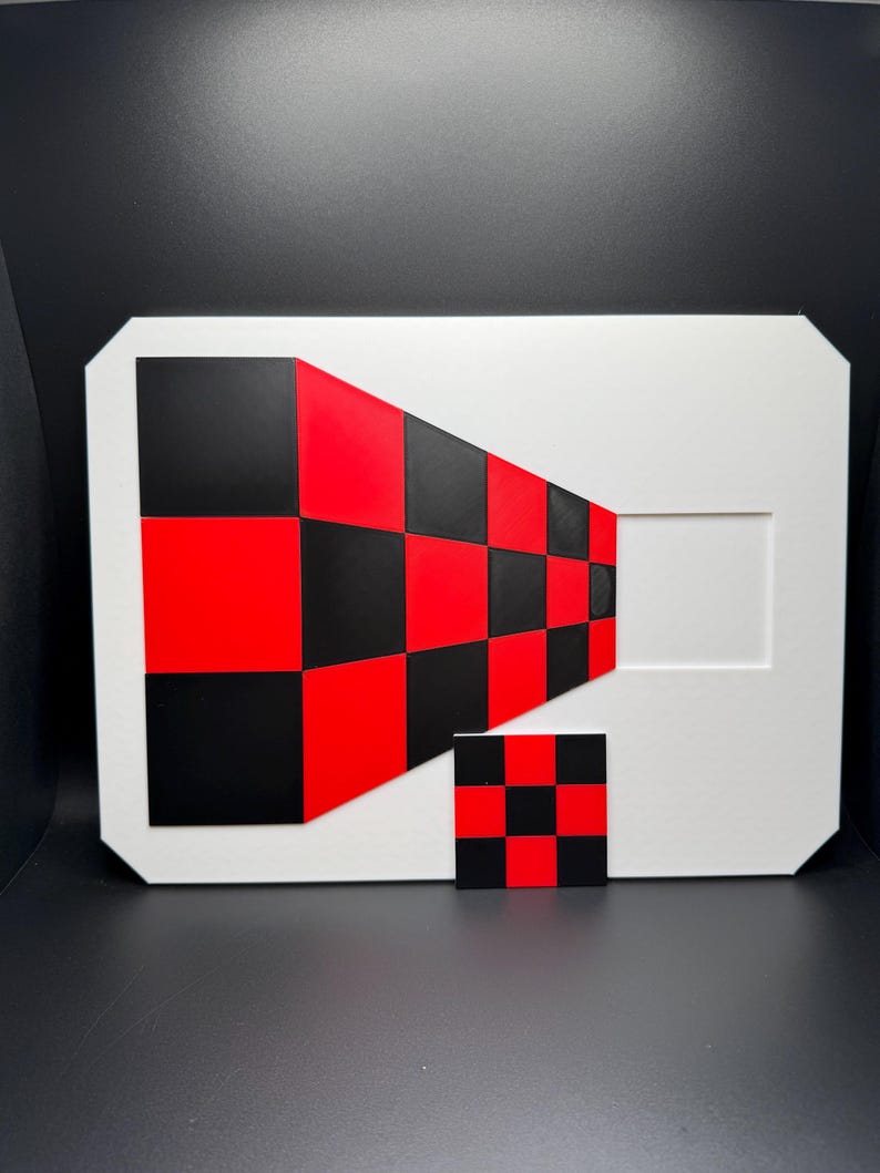 May include: A white rectangular art piece with a geometric design in red and black checkerboard pattern. A smaller, matching checkerboard square is in front of the larger piece. The artwork has a modern, abstract aesthetic.