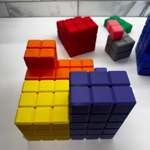 May include: A colorful set of building blocks made of plastic. The blocks are arranged in a tower formation, with the bottom layer being yellow, the middle layer being orange, and the top layer being purple. There are also smaller blocks in red, pink, and gray.