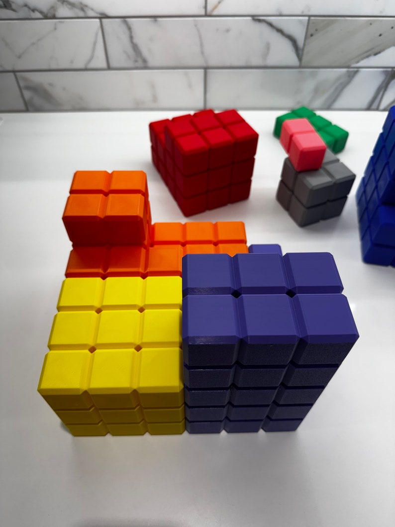 May include: A colorful set of building blocks made of plastic. The blocks are arranged in a tower formation, with the bottom layer being yellow, the middle layer being orange, and the top layer being purple. There are also smaller blocks in red, pink, and gray.