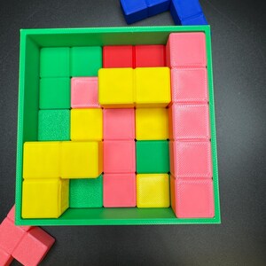 May include: A colorful puzzle game with a green square frame filled with various colored blocks. The blocks are red, pink, yellow, and green. A blue L-shaped block sits on top. The game is on a black surface.