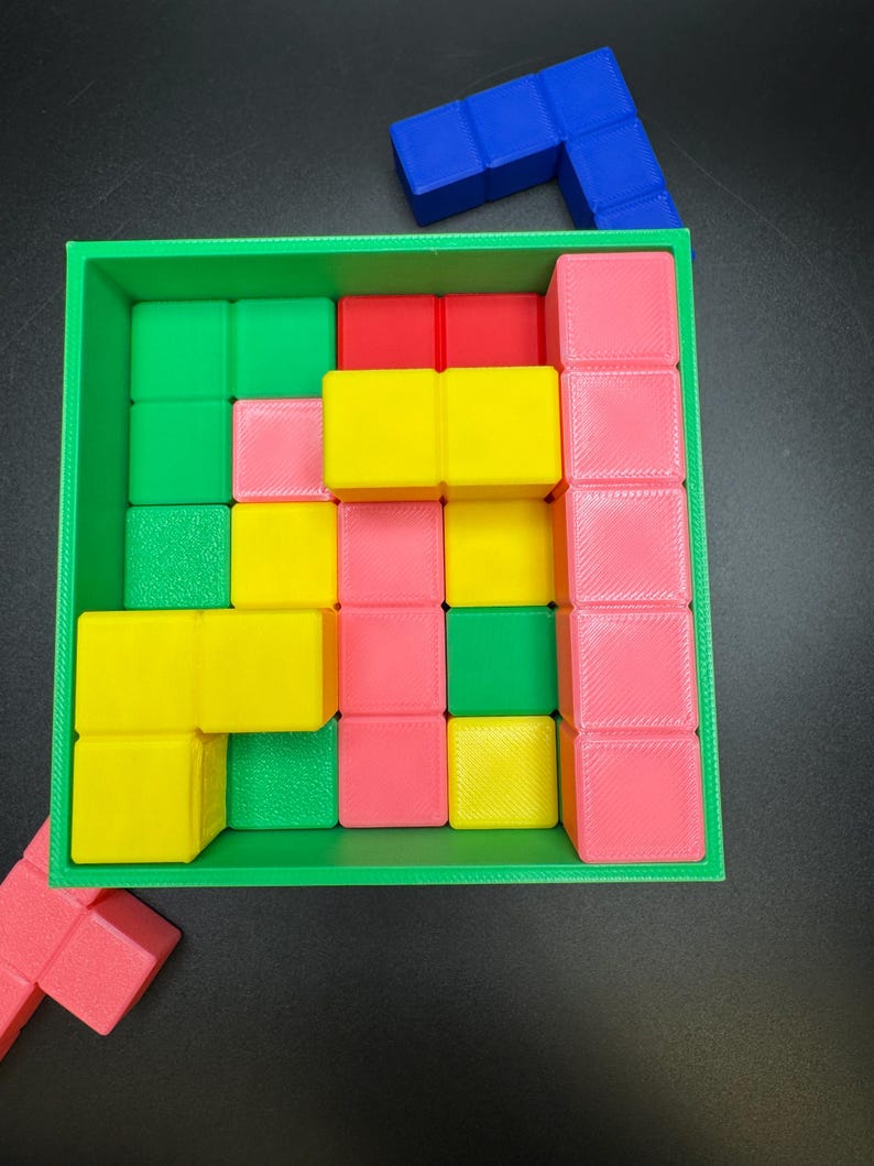 May include: A colorful puzzle game with a green square frame filled with various colored blocks. The blocks are red, pink, yellow, and green. A blue L-shaped block sits on top. The game is on a black surface.