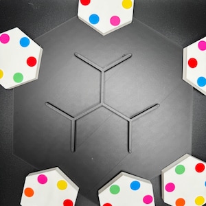 May include: Six hexagonal white tiles with colorful dots arranged in a circle around a black hexagonal base. The tiles are arranged in a pattern with the dots facing outward.