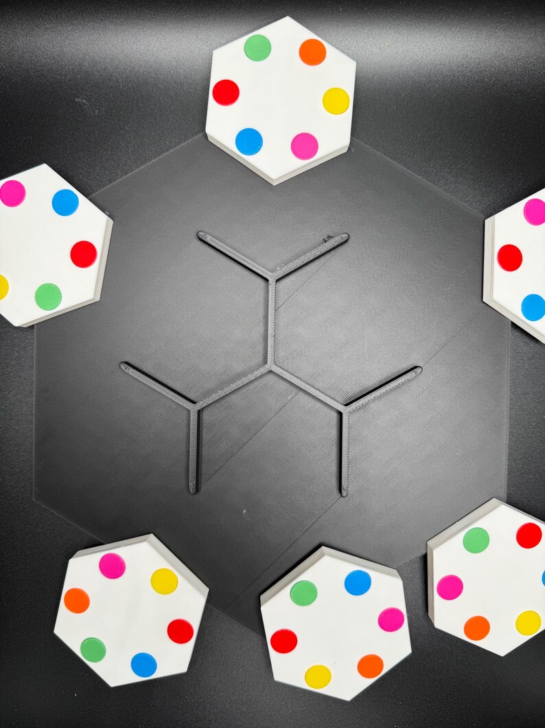May include: Six hexagonal white tiles with colorful dots arranged in a circle around a black hexagonal base. The tiles are arranged in a pattern with the dots facing outward.