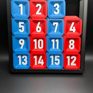 Pu&ograve; includere: Un puzzle scorrevole di 15 pezzi con tessere numerate in una cornice nera. Le tessere sono disposte in una griglia 4x4 con i numeri da 1 a 15. Le tessere sono blu e rosse.