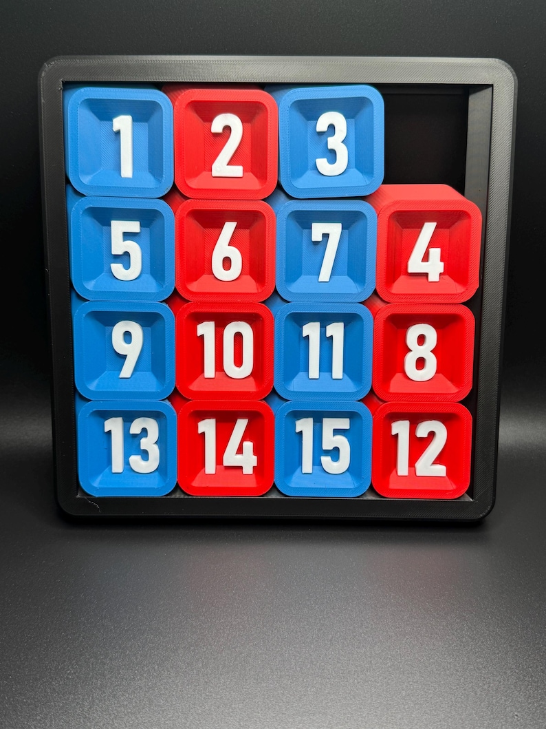 Pu&ograve; includere: Un puzzle scorrevole di 15 pezzi con tessere numerate in una cornice nera. Le tessere sono disposte in una griglia 4x4 con i numeri da 1 a 15. Le tessere sono blu e rosse.