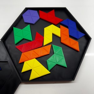 Op de afbeelding: Een zeshoekige puzzelbak met een zwarte basis en een verscheidenheid aan kleurrijke geometrische vormen. De vormen zijn gemaakt van plastic en omvatten een paarse zeshoek, rode driehoeken, gele driehoeken, groene driehoeken en blauwe driehoeken.