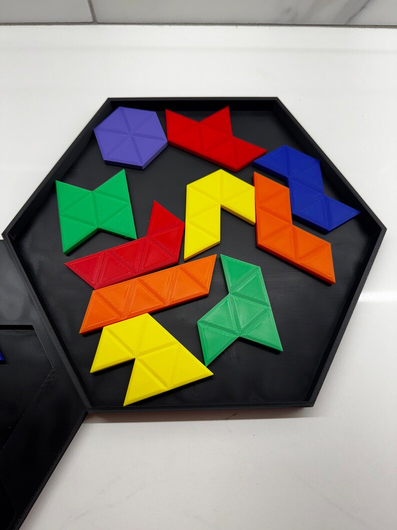 Op de afbeelding: Een zeshoekige puzzelbak met een zwarte basis en een verscheidenheid aan kleurrijke geometrische vormen. De vormen zijn gemaakt van plastic en omvatten een paarse zeshoek, rode driehoeken, gele driehoeken, groene driehoeken en blauwe driehoeken.