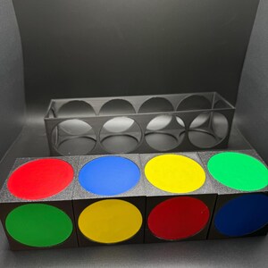 May include: A black storage unit with eight circular openings, and a matching set of eight black cubes. Each cube has a coloured, oval-shaped insert in red, blue, yellow, and green.