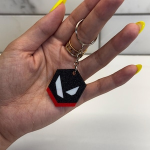 May include: Black and red hexagonal keychain with a white graphic design. The keychain is attached to a silver key ring. The keychain is held in a hand with yellow nail polish.