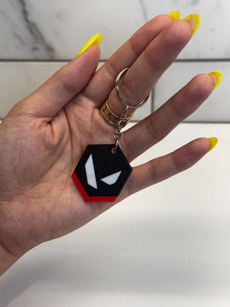 May include: Black and red hexagonal keychain with a white graphic design. The keychain is attached to a silver key ring. The keychain is held in a hand with yellow nail polish.