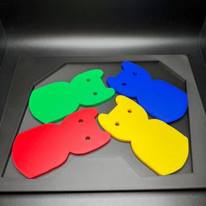 May include: Four colorful cat-shaped acrylic pieces in red, green, blue, and yellow. Each piece has two small holes and is set on a dark gray tray. The shapes are arranged in a playful, eye-catching display.