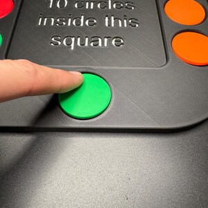 K&ouml;nnte beinhalten: Ein schwarzes Spielbrett mit bunten kreisf&ouml;rmigen Teilen. Auf dem Brett steht der Text "Place all 10 circles inside this square". Ein Finger ber&uuml;hrt einen gr&uuml;nen Kreis. Weitere Kreise sind rot, gelb und orange.