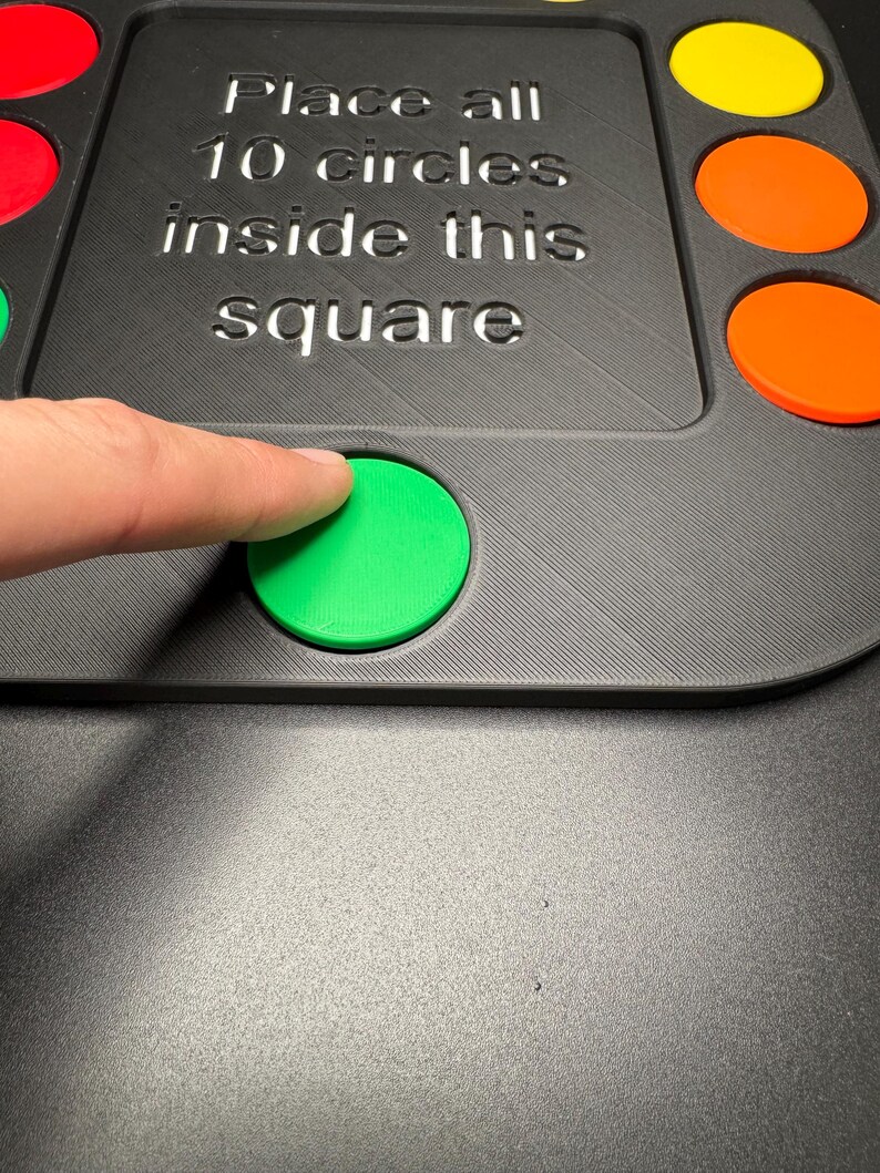 K&ouml;nnte beinhalten: Ein schwarzes Spielbrett mit bunten kreisf&ouml;rmigen Teilen. Auf dem Brett steht der Text "Place all 10 circles inside this square". Ein Finger ber&uuml;hrt einen gr&uuml;nen Kreis. Weitere Kreise sind rot, gelb und orange.