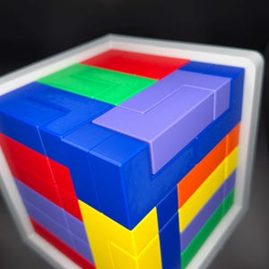 May include: A multi-colored 3D puzzle cube featuring interlocking geometric blocks in shades of red, blue, green, yellow, purple, and orange. The cube is encased in a white frame and displays a complex arrangement of various shapes on its surface.