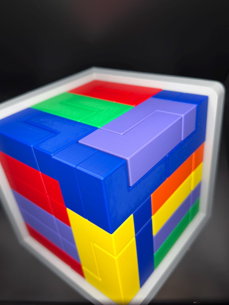 May include: A multi-colored 3D puzzle cube featuring interlocking geometric blocks in shades of red, blue, green, yellow, purple, and orange. The cube is encased in a white frame and displays a complex arrangement of various shapes on its surface.