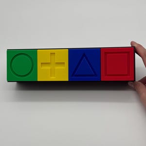 May include: A black rectangular box with four colored squares. The squares are green, yellow, blue, and red. Each square has a different symbol: a circle, a plus sign, a triangle, and a square.