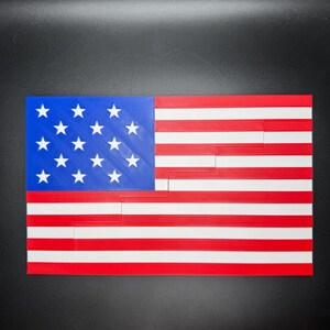 Puede incluir: Una bandera estadounidense impresa en 3D con un campo de estrellas azul y 13 rayas rojas y blancas. La bandera est&aacute; cortada en un patr&oacute;n escalonado.