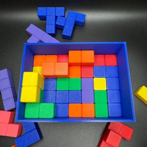 May include: A blue rectangular tray filled with colorful, geometric puzzle pieces. The pieces are various colors including blue, orange, red, green, yellow, and purple. Some pieces are outside the tray, scattered on a black surface.