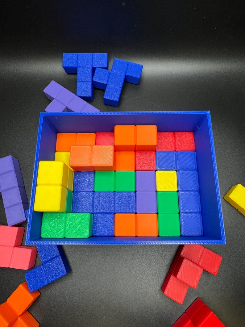 May include: A blue rectangular tray filled with colorful, geometric puzzle pieces. The pieces are various colors including blue, orange, red, green, yellow, and purple. Some pieces are outside the tray, scattered on a black surface.