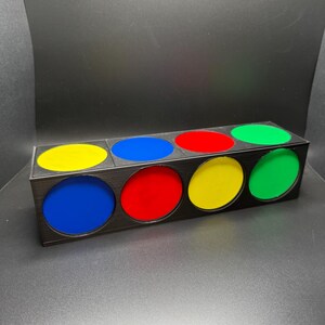 May include: A rectangular black box with six circular openings, each filled with a different colour: yellow, blue, red, yellow, and green. The box is made of a dark material and sits on a dark surface.