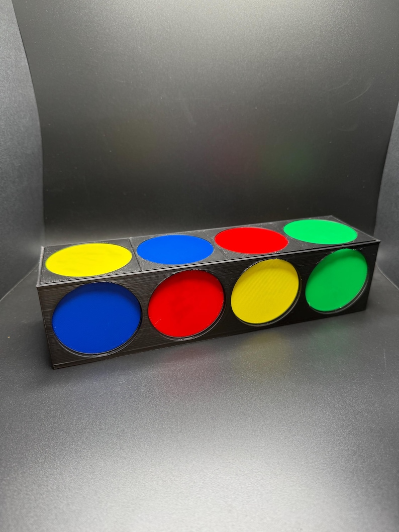 May include: A rectangular black box with six circular openings, each filled with a different colour: yellow, blue, red, yellow, and green. The box is made of a dark material and sits on a dark surface.