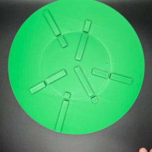 May include: A bright green, circular object with a series of rectangular protrusions arranged around the center. The object is set against a dark background. The design suggests a functional or decorative item.