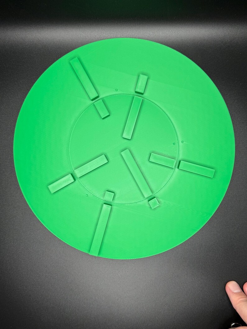 May include: A bright green, circular object with a series of rectangular protrusions arranged around the center. The object is set against a dark background. The design suggests a functional or decorative item.