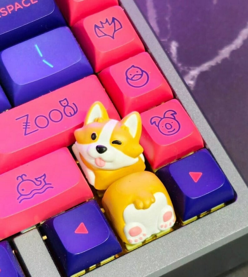 Op de afbeelding: Een aangepast toetsenbord met roze, paarse en blauwe toetsen. Een corgi-vormige toets knipoogt met zijn tong. Andere toetsen hebben dierlijke en geometrische ontwerpen.